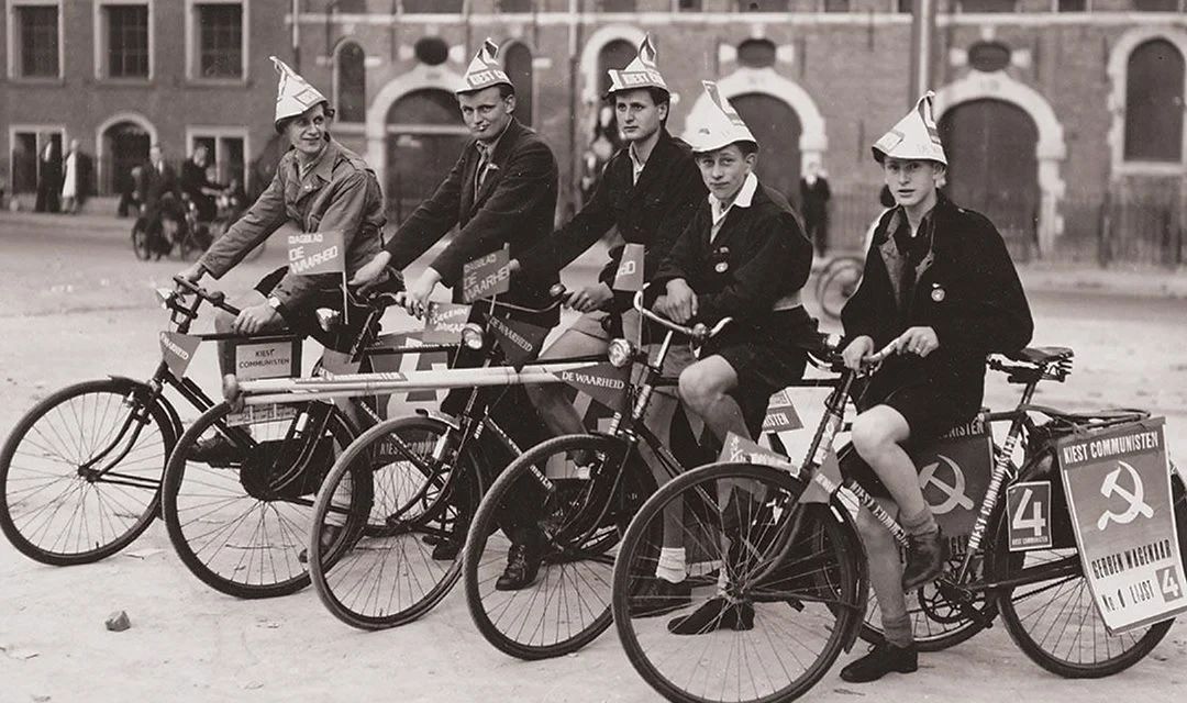 Five boys on bicycles with paper hats and signs Vote communist and Daily De Waarheid