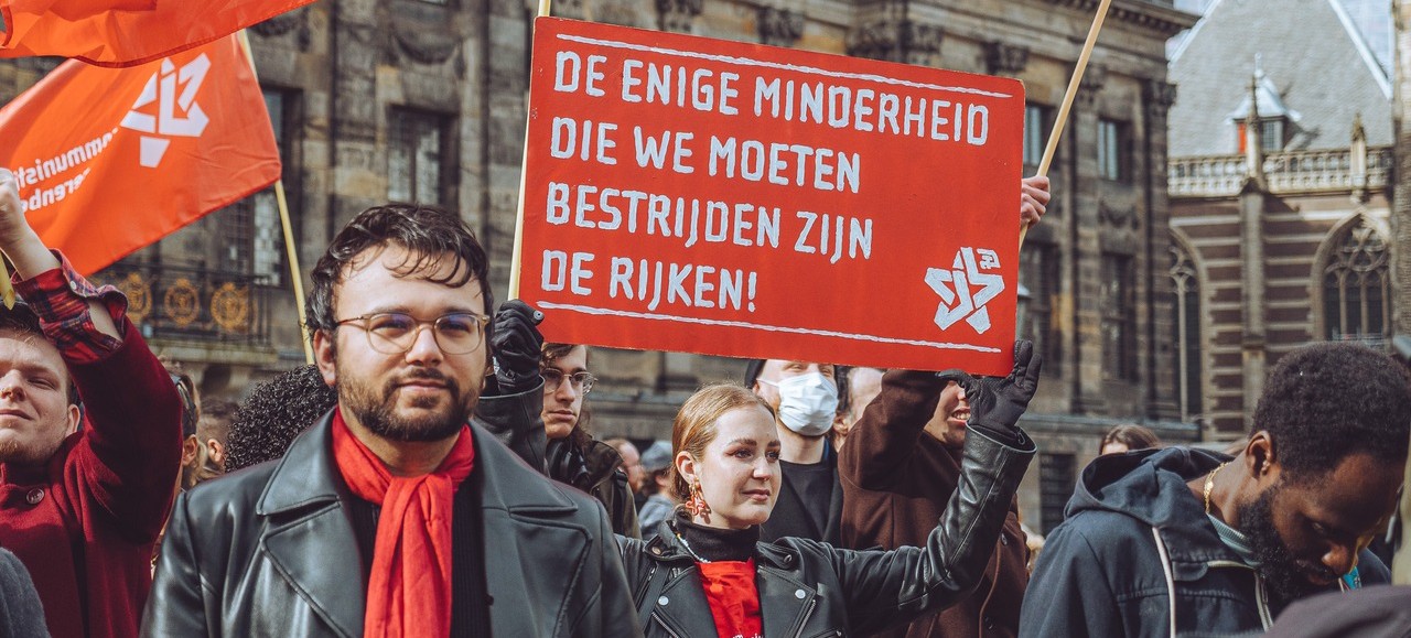 Demonstranten op de Dam met rode vlaggen en een bord: "De enige minderheid die we moeten bestrijden zijn de rijken!"