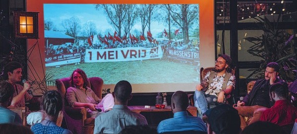 Gesprek in fauteuils voor publiek, op het scherm een foto van een 1 mei-demonstratie met rode vlaggen