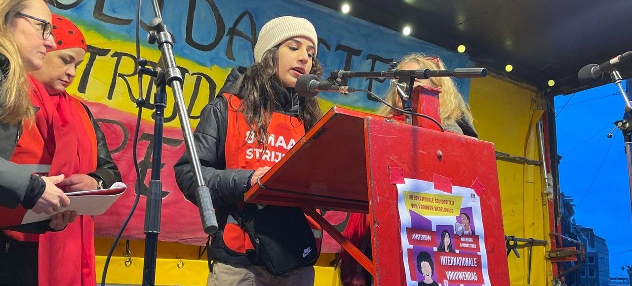 Een jonge vrouw spreekt achter een rood spreekgestoelte op het podium van de vrouwendagactie